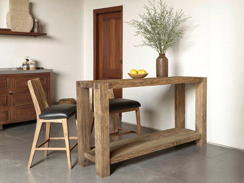 Peut inclure: Un &icirc;lot de cuisine en bois rustique avec une &eacute;tag&egrave;re inf&eacute;rieure, associ&eacute; &agrave; deux tabourets de bar en bois avec des si&egrave;ges en cuir noir. Un vase avec de la verdure et un bol de citrons sont pos&eacute;s sur l'&icirc;lot. La pi&egrave;ce a une porte en bois et un meuble.