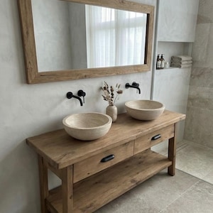 May include: A rustic bathroom vanity with two stone vessel sinks, black faucets, and a large wooden framed mirror. The vanity has two drawers and a lower shelf. A vase of dried flowers sits between the sinks.
