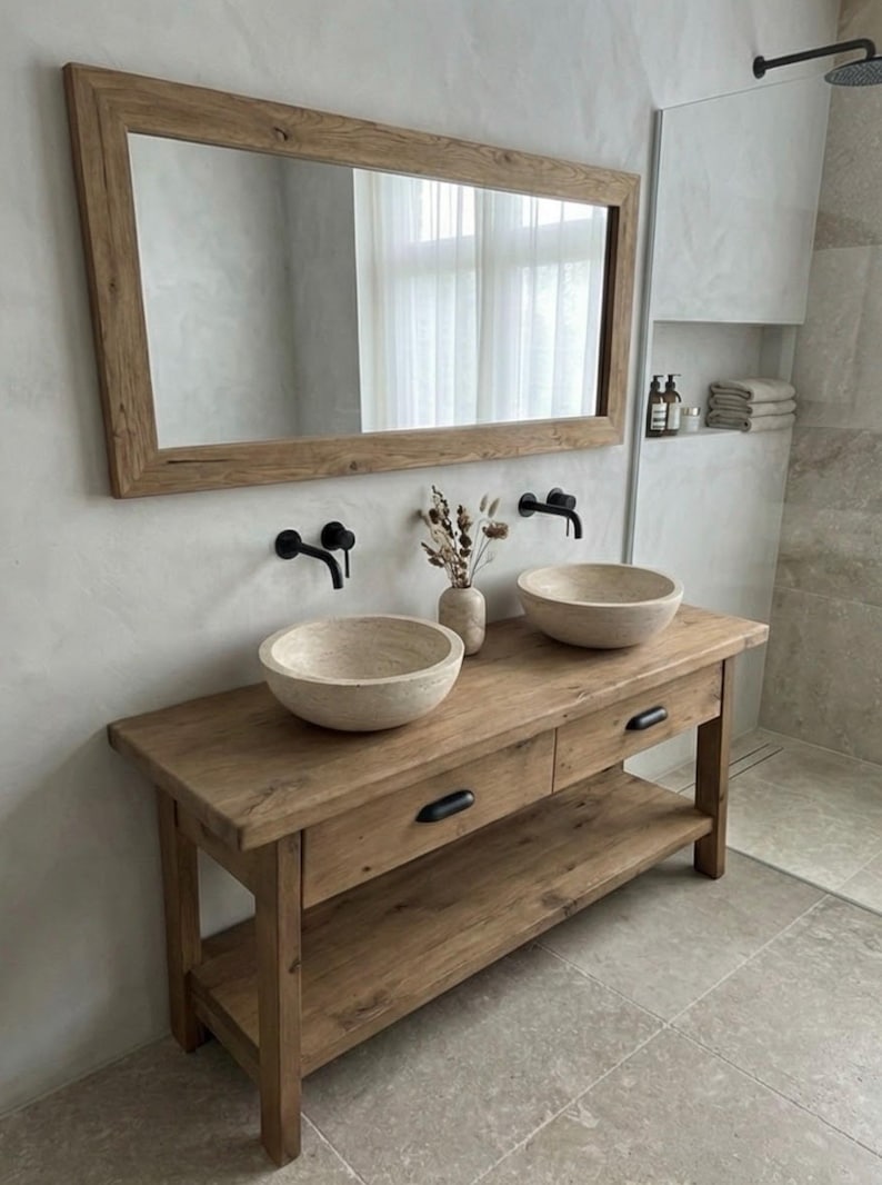 May include: A rustic bathroom vanity with two stone vessel sinks, black faucets, and a large wooden framed mirror. The vanity has two drawers and a lower shelf. A vase of dried flowers sits between the sinks.