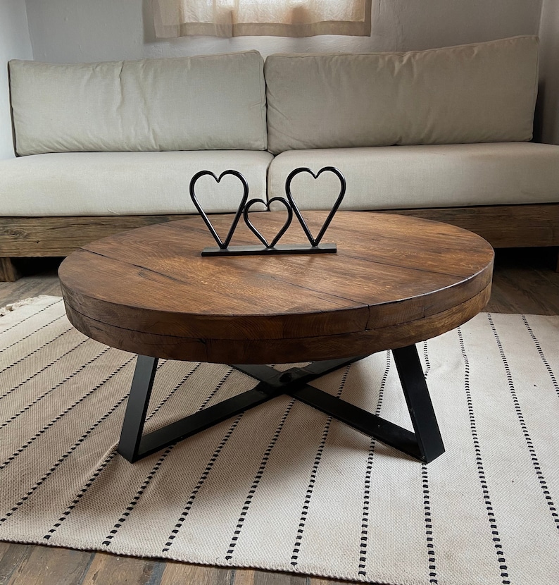 K&ouml;nnte beinhalten: Ein runder Holz-Couchtisch mit einem schwarzen Metallgestell. Der Tisch hat eine gealterte Holzoptik und steht auf einem wei&szlig;en Teppich mit schwarzen Streifen. Drei schwarze Metallskulpturen in Herzform befinden sich auf dem Tisch.