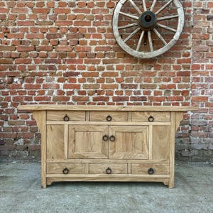May include: A light-colored wooden cabinet with multiple drawers and doors. The cabinet has black ring pulls and is set against a red brick wall with a wooden wagon wheel mounted above it.