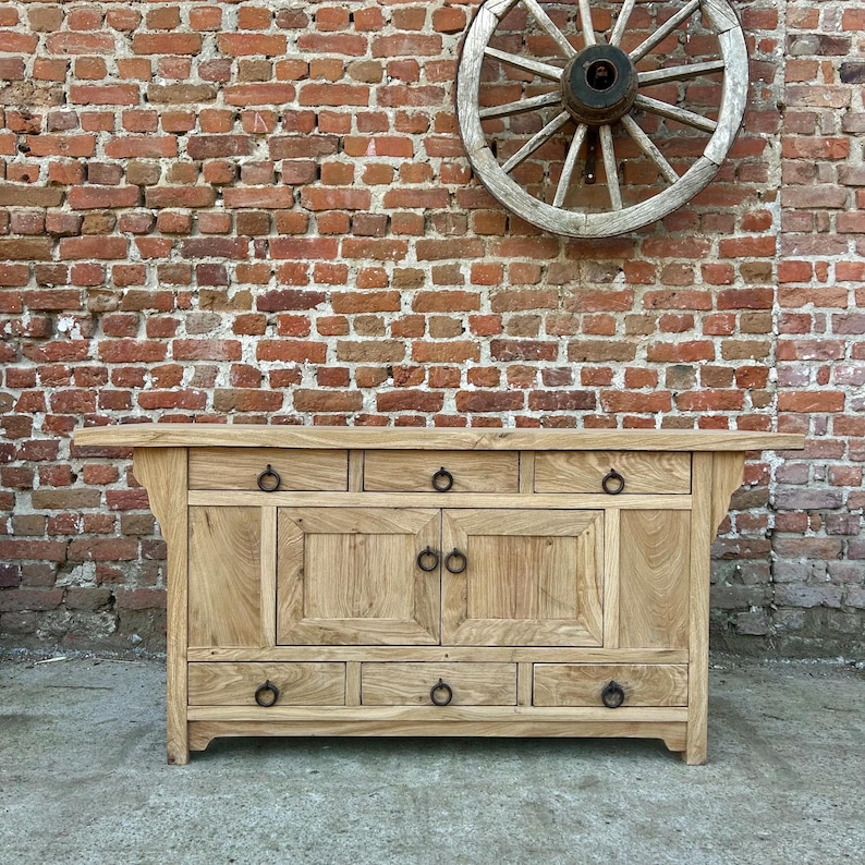 May include: A light-colored wooden cabinet with multiple drawers and doors. The cabinet has black ring pulls and is set against a red brick wall with a wooden wagon wheel mounted above it.