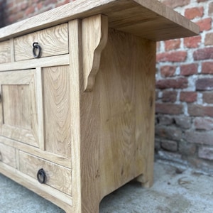 May include: A light oak wooden cabinet with drawers and a top shelf. The cabinet features decorative brackets and ring pulls on the drawers. The wood grain is visible, and the cabinet has a natural finish.