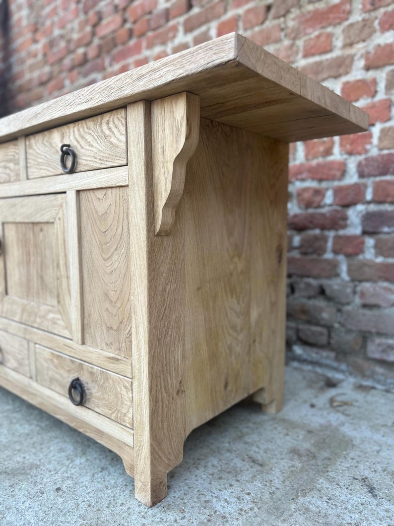 May include: A light oak wooden cabinet with drawers and a top shelf. The cabinet features decorative brackets and ring pulls on the drawers. The wood grain is visible, and the cabinet has a natural finish.