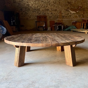 May include: A large, oval-shaped wooden coffee table with a rustic, weathered finish. The table has four thick, chunky legs that are slightly angled inwards. The table is made from reclaimed wood and has a natural, organic look.