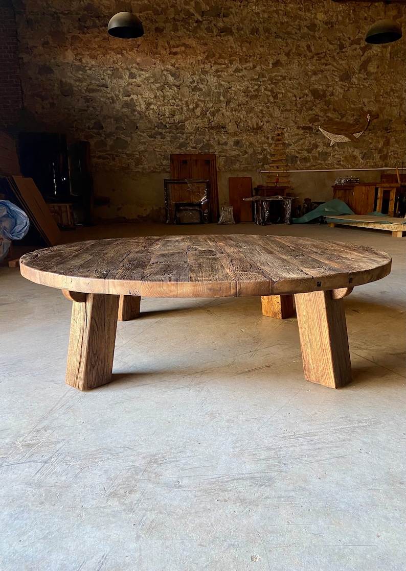 May include: A large, oval-shaped wooden coffee table with a rustic, weathered finish. The table has four thick, chunky legs that are slightly angled inwards. The table is made from reclaimed wood and has a natural, organic look.