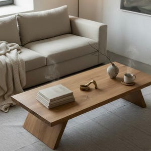 Puede incluir: Una mesa de centro de madera marr&oacute;n claro con patas anguladas. Una pila de libros, un objeto decorativo de lat&oacute;n, un jarr&oacute;n peque&ntilde;o y una taza sobre un posavasos est&aacute;n sobre la mesa. Un sof&aacute; beige y una manta color crema est&aacute;n en el fondo.
