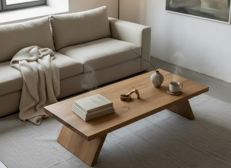 Puede incluir: Una mesa de centro de madera marr&oacute;n claro con patas anguladas. Una pila de libros, un objeto decorativo de lat&oacute;n, un jarr&oacute;n peque&ntilde;o y una taza sobre un posavasos est&aacute;n sobre la mesa. Un sof&aacute; beige y una manta color crema est&aacute;n en el fondo.