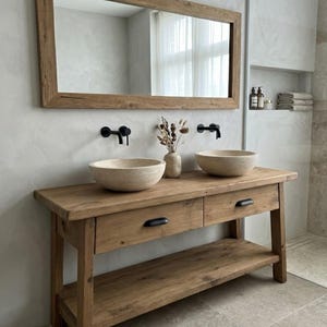 May include: A rustic wooden bathroom vanity with two round stone sinks and black faucets. A large wooden-framed mirror hangs above the vanity. The vanity has two drawers and a lower shelf. A vase with dried flowers sits between the sinks.
