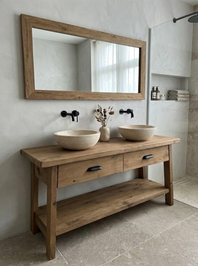 May include: A rustic wooden bathroom vanity with two round stone sinks and black faucets. A large wooden-framed mirror hangs above the vanity. The vanity has two drawers and a lower shelf. A vase with dried flowers sits between the sinks.