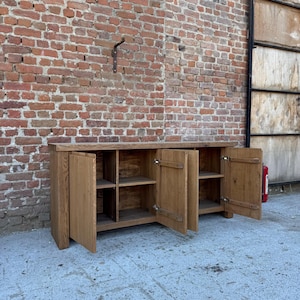 Peut inclure: Un long buffet rectangulaire en bois avec une finition brun clair naturelle. Le meuble poss&egrave;de quatre portes de placard, dont deux sont ouvertes pour r&eacute;v&eacute;ler des &eacute;tag&egrave;res int&eacute;rieures. Il est construit avec des panneaux de bois massif &eacute;pais.