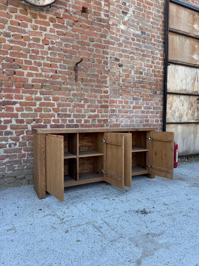 Peut inclure: Un long buffet rectangulaire en bois avec une finition brun clair naturelle. Le meuble poss&egrave;de quatre portes de placard, dont deux sont ouvertes pour r&eacute;v&eacute;ler des &eacute;tag&egrave;res int&eacute;rieures. Il est construit avec des panneaux de bois massif &eacute;pais.