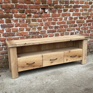 May include: A light oak wood TV stand with three drawers and a lower shelf. The stand has a natural finish and black metal handles. The stand is 60 inches long, 18 inches deep, and 20 inches high.