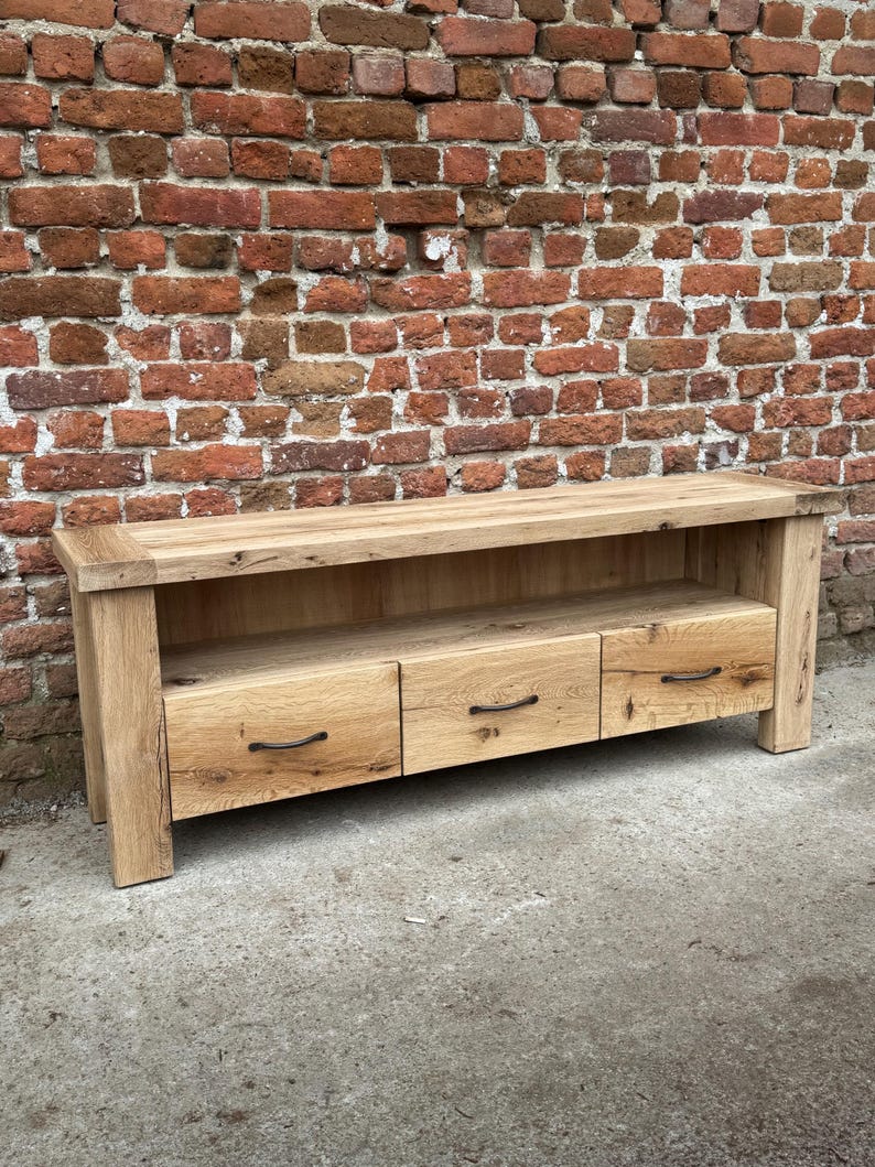 May include: A light oak wood TV stand with three drawers and a lower shelf. The stand has a natural finish and black metal handles. The stand is 60 inches long, 18 inches deep, and 20 inches high.