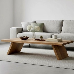 Puede incluir: Una mesa de centro rectangular de madera clara con patas anguladas. La mesa tiene un acabado de veta de madera natural. Un sof&aacute; y una alfombra grises est&aacute;n en el fondo. Incluye un cuenco, libros y una peque&ntilde;a planta.