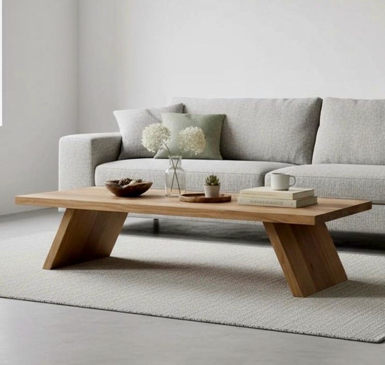 Puede incluir: Una mesa de centro rectangular de madera clara con patas anguladas. La mesa tiene un acabado de veta de madera natural. Un sof&aacute; y una alfombra grises est&aacute;n en el fondo. Incluye un cuenco, libros y una peque&ntilde;a planta.