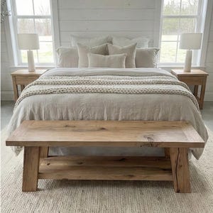 May include: A bedroom scene featuring a bed with neutral-toned bedding and pillows. A chunky knit throw adds texture. Two bedside tables with lamps flank the bed. A rustic wooden bench sits at the foot of the bed, and a woven rug covers the floor.