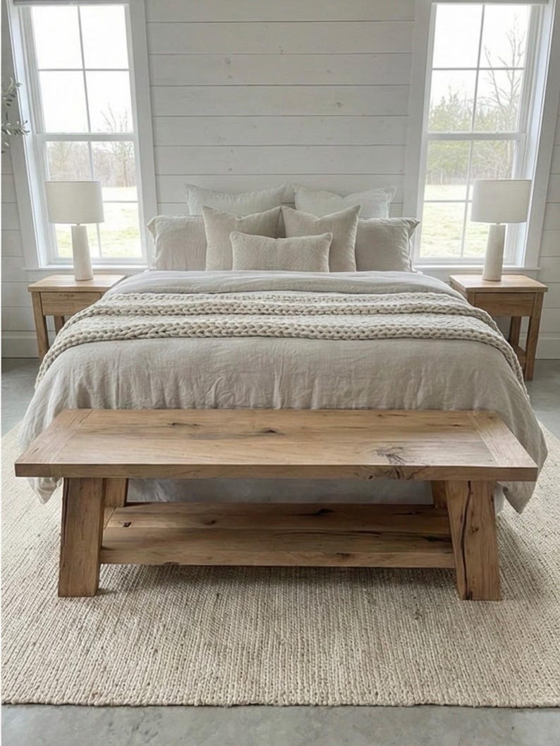 May include: A bedroom scene featuring a bed with neutral-toned bedding and pillows. A chunky knit throw adds texture. Two bedside tables with lamps flank the bed. A rustic wooden bench sits at the foot of the bed, and a woven rug covers the floor.