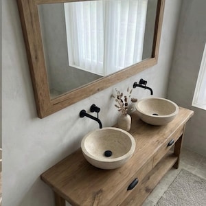 May include: A rustic bathroom vanity with a large wooden-framed mirror. The vanity features two stone vessel sinks, black faucets, and a wooden cabinet with drawers. A vase with dried flowers sits between the sinks.