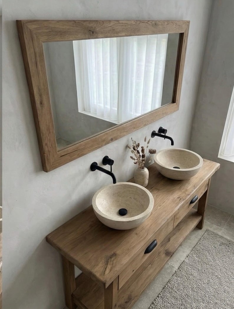May include: A rustic bathroom vanity with a large wooden-framed mirror. The vanity features two stone vessel sinks, black faucets, and a wooden cabinet with drawers. A vase with dried flowers sits between the sinks.