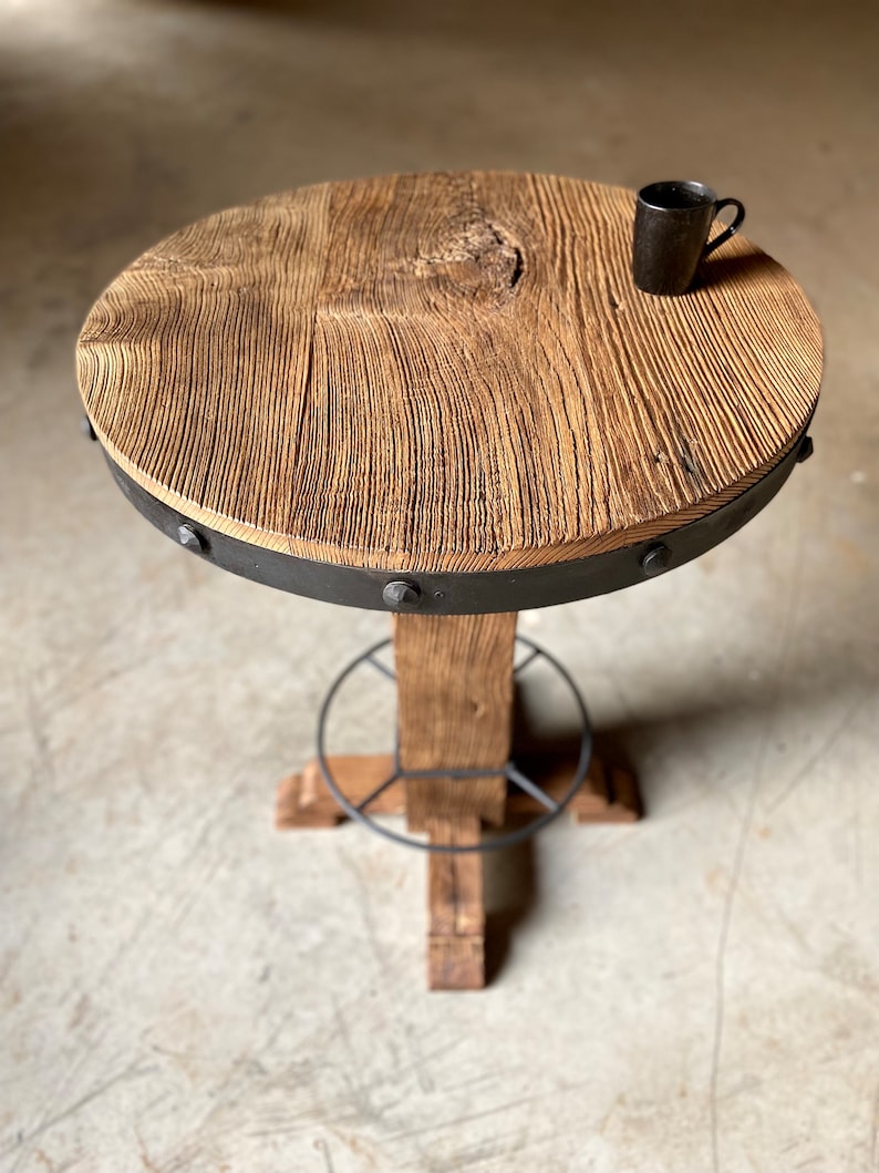 May include: A round wooden table with a metal base and a black metal ring around the top. The table has a rustic look and is made of reclaimed wood.