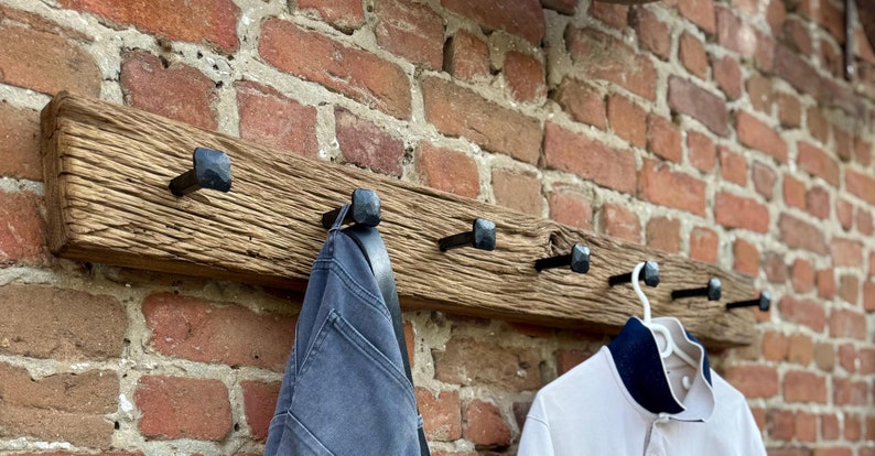 May include: A rustic wooden coat rack with five black metal hooks. The rack is mounted on a brick wall and has a weathered, distressed finish. A blue denim jacket and a white polo shirt are hanging on the hooks.
