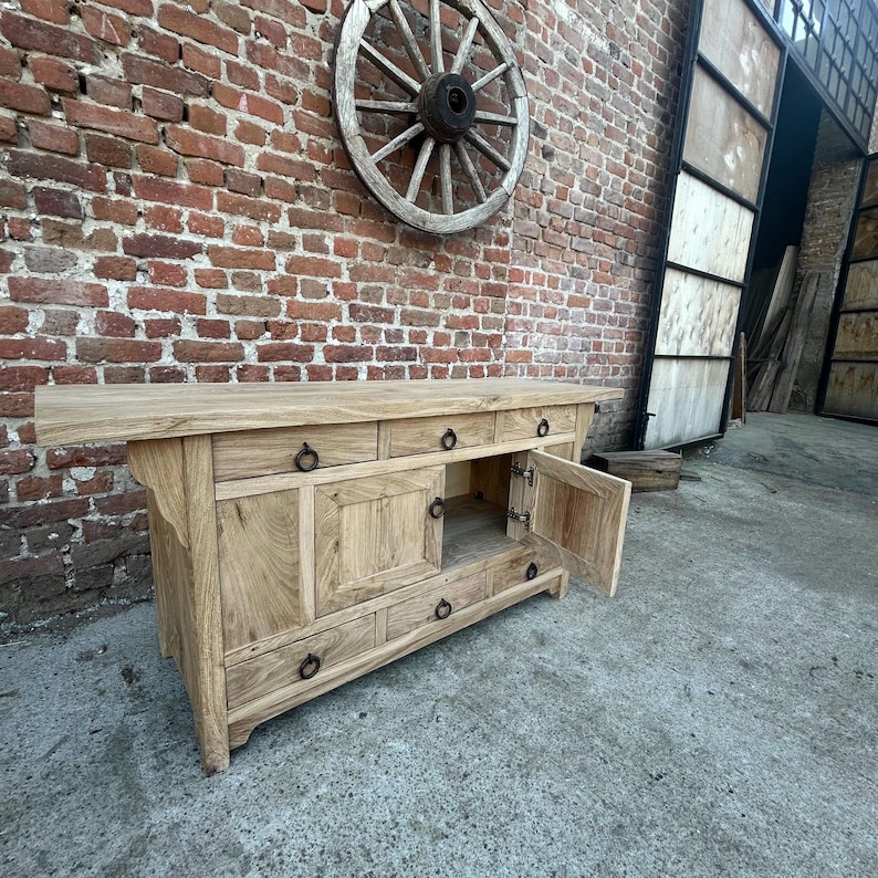 May include: A light-colored wooden sideboard with multiple drawers and cabinet doors, some open. The sideboard has black ring pulls and is set against a brick wall with a wooden wagon wheel. The cabinet is made of light-colored wood.