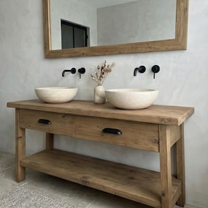 May include: A rustic wooden bathroom vanity with two drawers and a lower shelf. Two stone vessel sinks sit atop the vanity, with black faucets mounted on the wall. A large wooden-framed mirror hangs above the vanity, and a vase of dried flowers sits between the sinks.