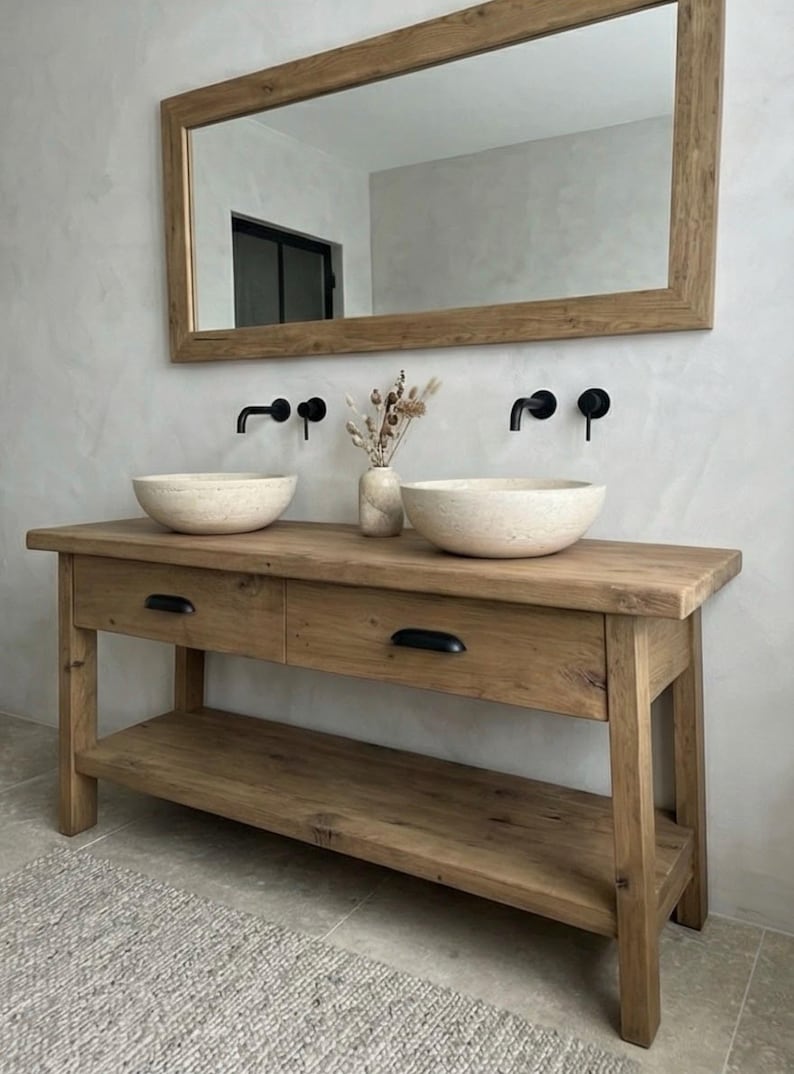 May include: A rustic wooden bathroom vanity with two drawers and a lower shelf. Two stone vessel sinks sit atop the vanity, with black faucets mounted on the wall. A large wooden-framed mirror hangs above the vanity, and a vase of dried flowers sits between the sinks.