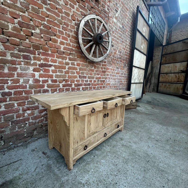 May include: A light-colored wooden cabinet with multiple drawers and doors. The cabinet has a rustic design with metal hardware. A wooden wagon wheel hangs on the brick wall behind the cabinet.