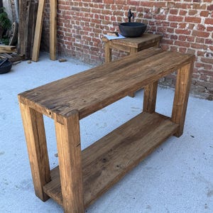 Peut inclure: Une console en bois rustique avec une &eacute;tag&egrave;re inf&eacute;rieure. La table est en bois brun clair et de forme rectangulaire. Elle est plac&eacute;e devant un mur de briques. Id&eacute;ale pour un salon ou une entr&eacute;e. Dimensions en centim&egrave;tres.