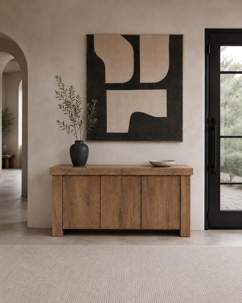 Peut inclure: Un buffet rectangulaire en bois dot&eacute; de trois portes verticales et d'un plateau &eacute;pais. Sur le buffet se trouvent un vase noir contenant des branches feuillues vertes et un petit bol peu profond de couleur claire. Une grande &oelig;uvre murale carr&eacute;e est suspendue au-dessus.