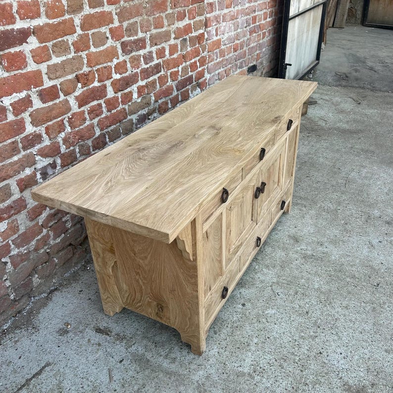 May include: A light-colored wooden cabinet with a large, flat top and multiple drawers and doors. The cabinet has dark metal hardware and a natural wood grain finish. The cabinet is made of solid wood and has a rustic, handcrafted appearance.
