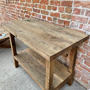 May include: A rustic wooden table with a shelf. The table is made of reclaimed wood and has a natural finish. It is perfect for a farmhouse or industrial style home.
