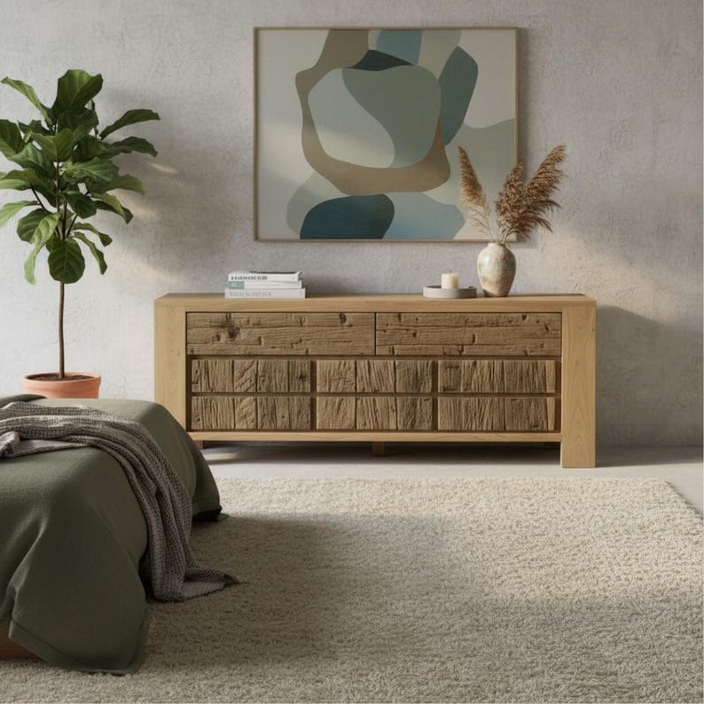 May include: A light oak wood dresser with a textured front, featuring two sets of drawers. Above the dresser is a framed abstract painting with neutral colors. A potted plant and a vase with dried flowers are also visible, creating a warm, inviting interior design.