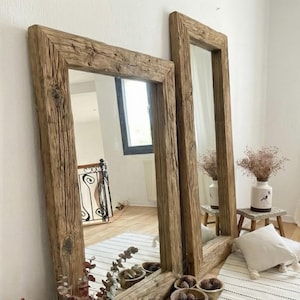May include: Two large rectangular mirrors with thick, rustic wooden frames. The frames have a natural, weathered appearance. The mirrors are leaning against a white wall, reflecting the room. The style is farmhouse or rustic.