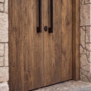 May include: A set of double doors made from weathered brown wood with a prominent grain pattern. The doors feature long, vertical, matte black metal handles and a central circular keyhole escutcheon positioned between the two door panels.