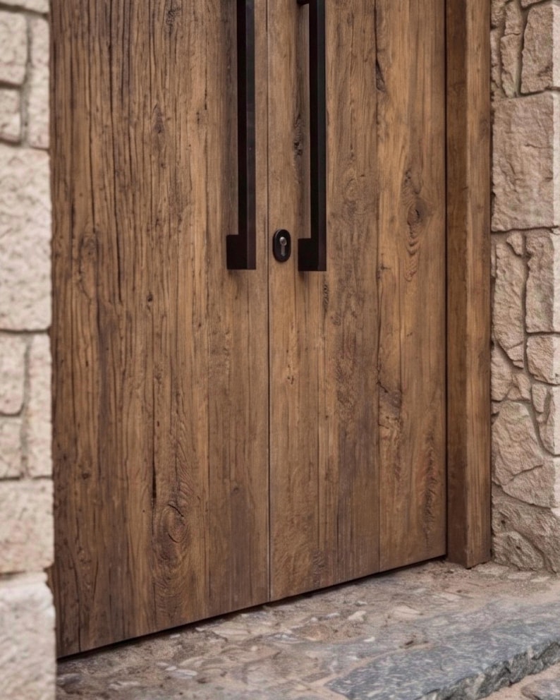 May include: A set of double doors made from weathered brown wood with a prominent grain pattern. The doors feature long, vertical, matte black metal handles and a central circular keyhole escutcheon positioned between the two door panels.