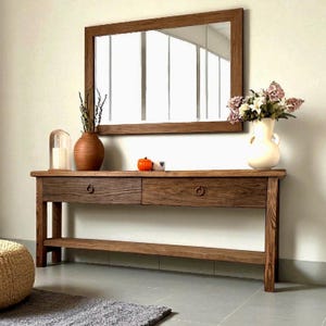 May include: A wooden console table with two drawers and a lower shelf, topped with decorative items. A large rectangular mirror hangs above the table. The table and mirror have a warm, brown finish. A vase of flowers and other decor are on the table.