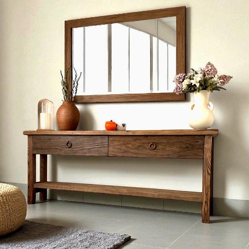 May include: A wooden console table with two drawers and a lower shelf, topped with decorative items. A large rectangular mirror hangs above the table. The table and mirror have a warm, brown finish. A vase of flowers and other decor are on the table.