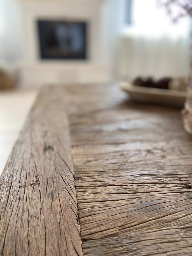 May include: Close-up of a weathered, rustic wooden table. The wood has a natural, aged appearance with visible grain and cracks. The table's surface is a warm, brown tone, suggesting it's made from reclaimed or distressed wood. The focus is on the texture and detail of the wood.