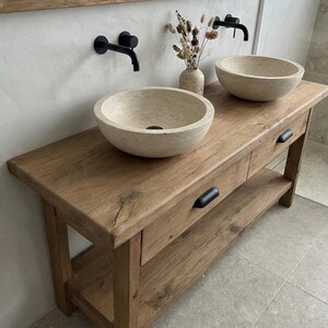 May include: A rustic wooden bathroom vanity with two stone vessel sinks and black faucets. The vanity has two drawers and a lower shelf. A mirror with a wooden frame hangs above the vanity. The overall design is natural and minimalist.
