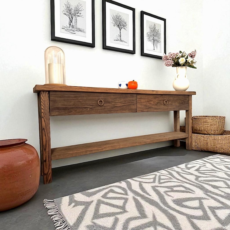 May include: A wooden console table with two drawers and a lower shelf. The table is made of dark wood and has two round drawer pulls. Above the table are three framed tree prints. A vase of flowers, two baskets, and a decorative rug are also visible.