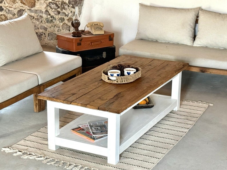 May include: White coffee table with a wooden top and a lower shelf. The table has a woven tray with two blue and white cups and pine cones on top. The table is on a white and brown striped rug.
