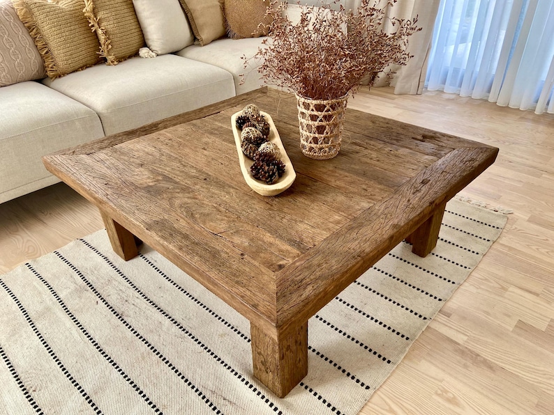May include: A rustic, square wooden coffee table with a distressed finish. The table is supported by thick legs and features a decorative wooden bowl with pinecones and a woven vase with dried flowers. A cream-colored sofa and rug are in the background.