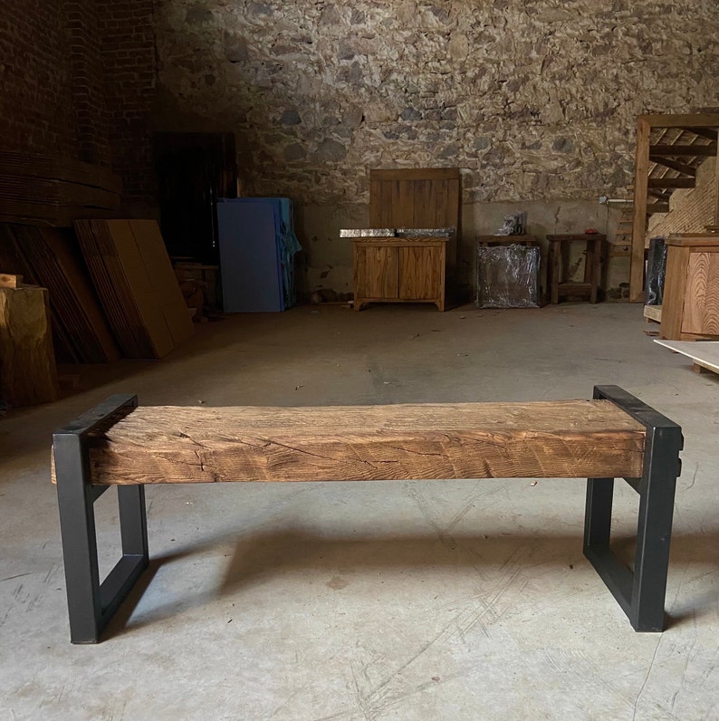 May include: A rustic wooden bench with a thick, weathered top and black metal legs. The bench is in a large, empty room with exposed brick walls.