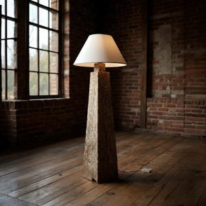 May include: A rustic floor lamp with a square, weathered wooden base and a white lampshade. The lamp stands tall in a room with exposed brick walls and a large window. The lamp is approximately 60 inches tall.