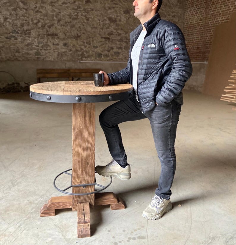 May include: A rustic wooden table with a black metal rim and a footrest. The table has a distressed wood finish and a round top.