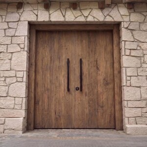May include: A set of double wooden doors with a natural brown grain finish, featuring two long, vertical black metal handles and a central circular lock. The doors are framed by a wall constructed from light-colored, irregular cut stone blocks.