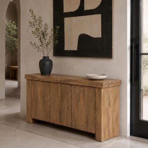 Peut inclure: Une console rectangulaire en bois avec un plateau &eacute;pais et quatre portes verticales pr&eacute;sentant une finition au grain naturel. Un vase en c&eacute;ramique gris anthracite contenant des branches feuillues et un petit bol en pierre mouchet&eacute;e.
